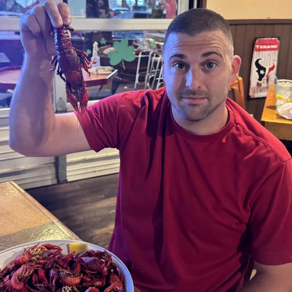 Man in red shirt holding up a crawfish, with a bucket of crawfish on the table.