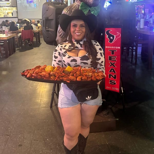 Smiling woman in cow print top and cowboy hat holding a large tray of crawfish and corn.