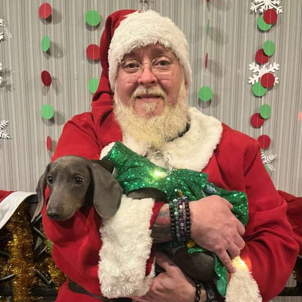 A man dressed as Santa Claus holds a dachshund wearing a green sequined outfit.