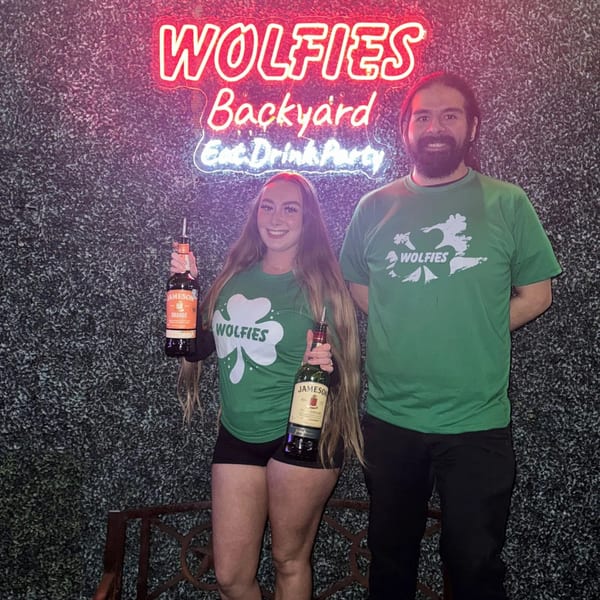 Two Wolfies staff members in green shirts holding Jameson bottles under a neon sign.