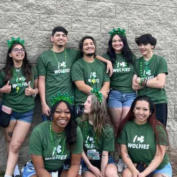 Wolfies staff in green shirts and St. Patrick's Day headbands pose against a stone wall.