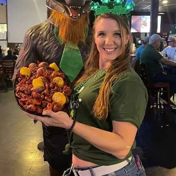 A woman in a green shirt and shamrock headband holds a large platter of crawfish and corn.