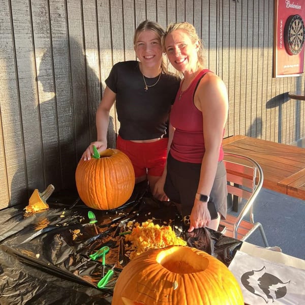 Two women smiling while carving pumpkins outdoors at Wolfies Restaurant and Sports Bar.
